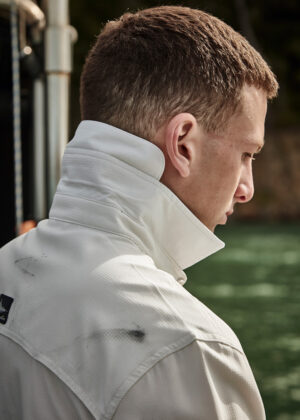 Profile view of a man with short brown hair wearing a white high-collared jacket, standing near water outdoors