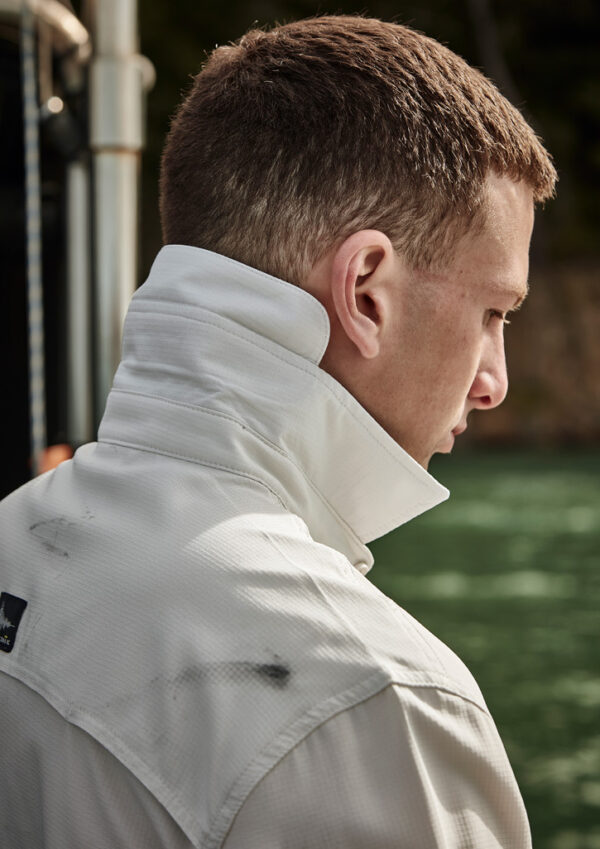 Profile view of a man with short brown hair wearing a white high-collared jacket, standing near water outdoors