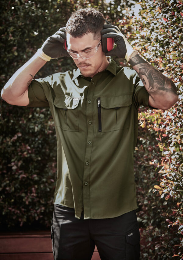 Man outdoors wearing safety gloves, goggles, and earmuffs, adjusting them over a green work shirt.