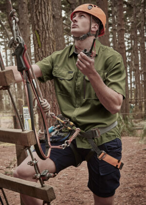 Man wearing a helmet and harness adjusts ropes on a forest ropes course obstacle, looking upward.