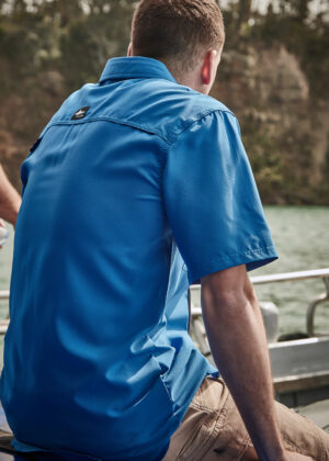 Back view of a man in a blue short-sleeve shirt seated on a boat by the water, facing away from the camera.