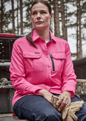 Woman in a bright pink work shirt sits outdoors, hands resting on her lap with gloves, trees in the background.