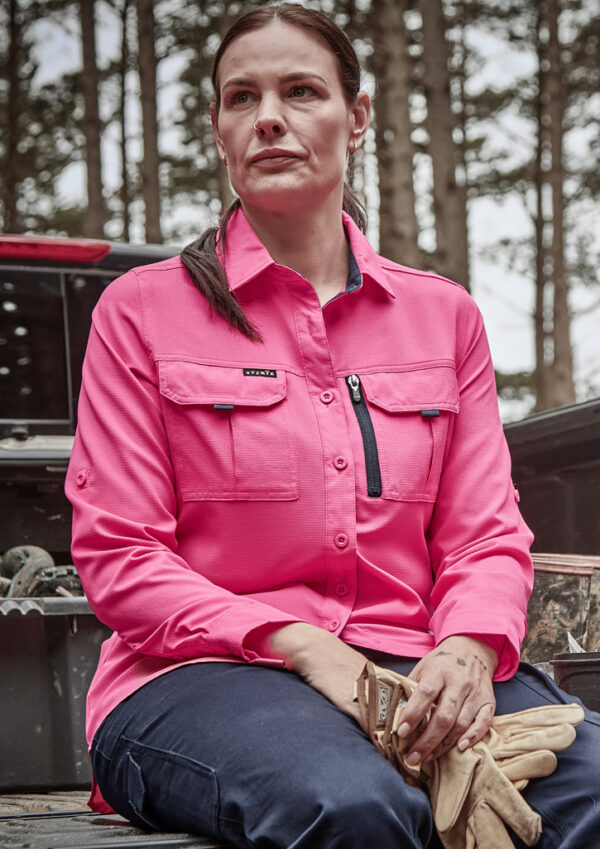 Woman in a bright pink work shirt sits outdoors, hands resting on her lap with gloves, trees in the background.