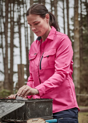 Woman in a pink button-up shirt working outdoors in a forest, opening a black toolbox and handling tools inside.