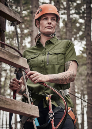 Woman wearing an orange helmet in a green utility shirt, adjusting climbing gear on a ropes course outdoors.