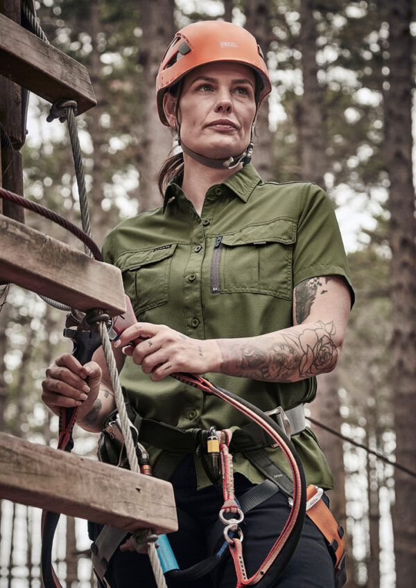 Woman wearing an orange helmet in a green utility shirt, adjusting climbing gear on a ropes course outdoors.