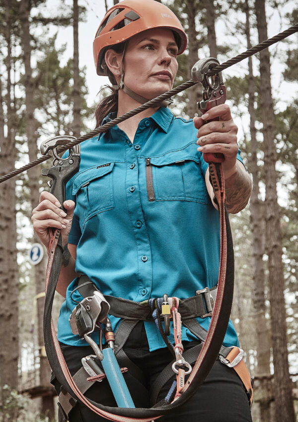 Woman in an orange helmet wearing a teal shirt, handling climbing ropes and carabiners on a safety harness in a forest setting.