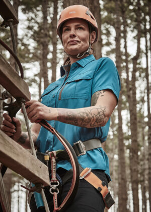 Climber wearing an orange helmet and turquoise shirt adjusts a rope on a tree‑top rope course in a forest.